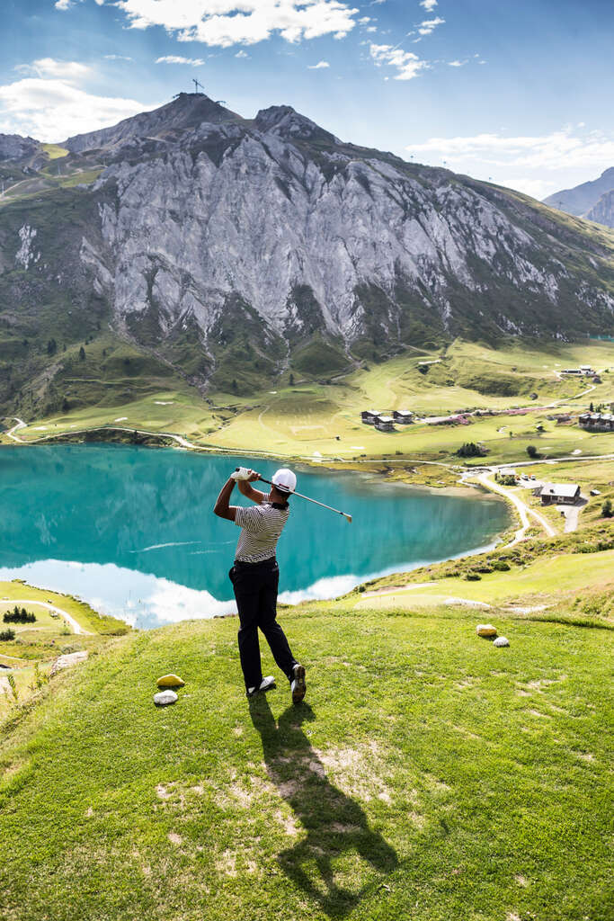 A golfer following through on a tee box above an alpine lake, with limestone peaks behind